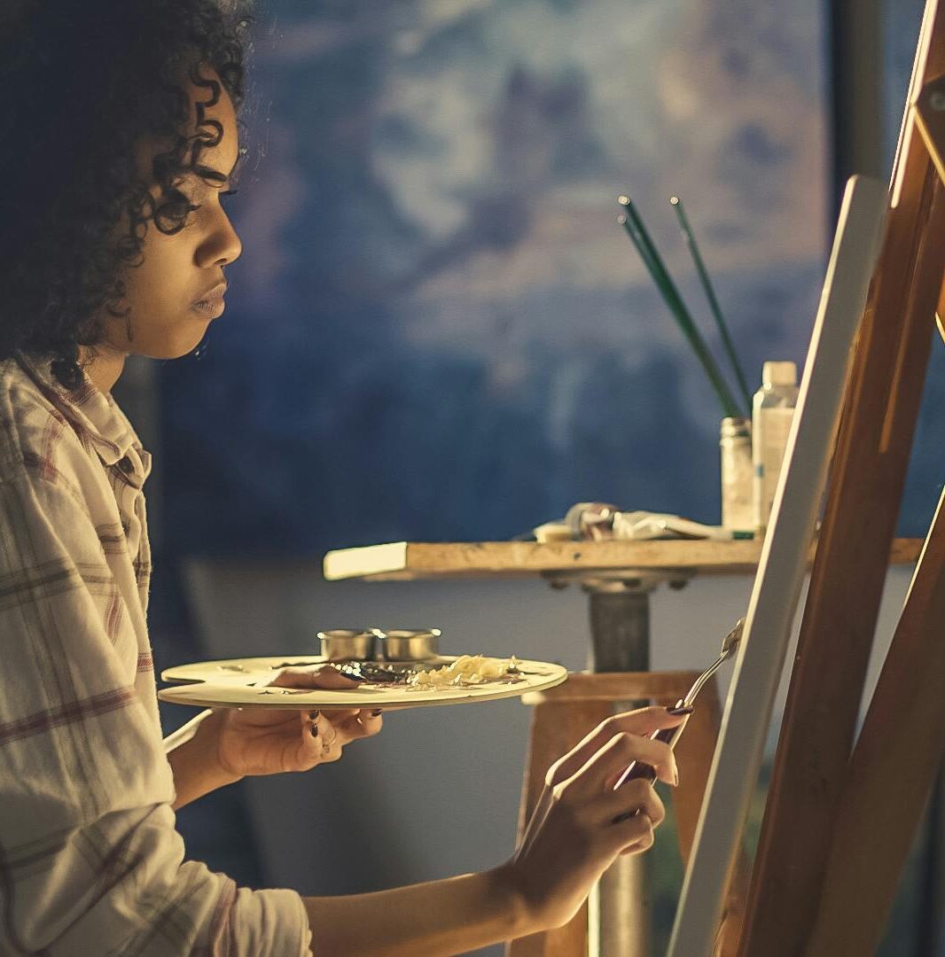 Side view of a young woman artist painting on canvas in a cozy studio setting.